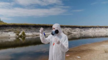 Koruyucu giysili ve solunum cihazı giyen adam, kirli nehirden analiz için aldığı test tüpündeki suyun kimyasal reaksiyonuna dikkat çekti. Bilim adamı örnek lerini in vitro olarak alır. Ekolojik felaket. 4k