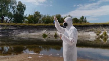 Koruyucu giysili ve solunum cihazı giyen adam, kirli nehirden analiz için aldığı test tüpündeki suyun kimyasal reaksiyonuna dikkat çekti. Bilim adamı örnek lerini in vitro olarak alır. Ekolojik felaket. 4k