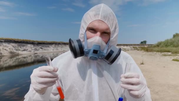 防護服を着た男とインビトロの人工呼吸器は 人々がプラスチック製のゴミを残し カメラに見える乾燥した川の隣のビーチに立っています 生態学的災害人為的影響4K — ストック動画