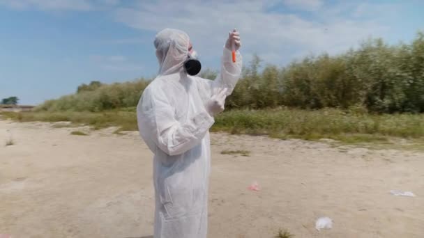 防護服と人工呼吸器を着た男性は 汚染された川からの分析のために取った試験管内の水の化学反応を観察します 科学者はインビトロでサンプルを採取します 生態学的災害4K — ストック動画