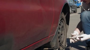 Beyaz eldivenli tecrübesiz bir adam kırık ve kirli kırmızı bir arabayı çaldı ve tekerleği sökmeye çalışıyor. Genç adam dışarıda araba tamir ediyor. Araba yolda bozuldu. Close. 4k görüntü.