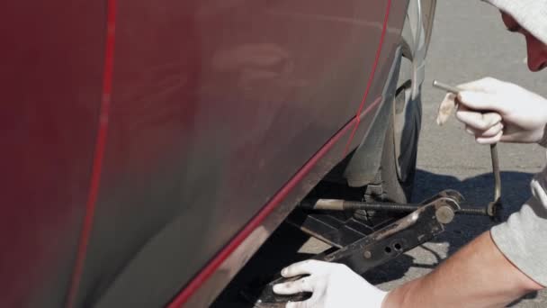 白い手袋を着た経験の浅い男は 壊れて汚れた赤い車をジャックしようとしています 屋外で自動を修理する若い男 その車は道路で故障した クローズ アップ 4Kフッテージ — ストック動画