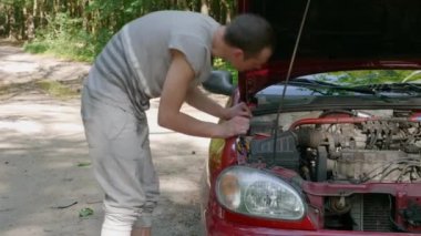 Deneyimsiz adam kırık arabanın açık kaputuna yaklaşır ve arıza nedeni ne olduğunu anlamaya çalışır. Genç adam dışarıda oto tamir ediyor. Otomobil ormanda bozuldu. 4k görüntü.