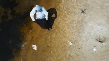 Koruyucu giysili bir adamın havadan görünümü ve kuru ve kirli bir nehrin kıyısında plastik çöp toplayan bir solunum cihazı. Ekolojik felaket. Antropojenik etki. 4k drone görüntüleri.
