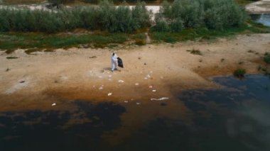 Kuru ve kirli bir nehrin kıyısında plastik çöp toplayan koruyucu giysili bir adamın havadan görünümü. Ekolojik felaket. Antropojenik etki. 4k drone görüntüleri.