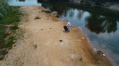 Kuru ve kirli bir nehrin kıyısında plastik çöp toplayan koruyucu giysili bir adamın havadan görünümü. Ekolojik felaket. Antropojenik etki. 4k drone görüntüleri.