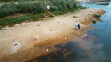 Kuru ve kirli bir nehrin kıyısında plastik çöp toplayan koruyucu giysili bir adamın havadan görünümü. Ekolojik felaket. Antropojenik etki. 4k drone görüntüleri.