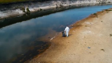 Kuru ve kirli bir nehrin kıyısında plastik çöp toplayan koruyucu giysili bir adamın havadan görünümü. Ekolojik felaket. Antropojenik etki. 4k drone görüntüleri.