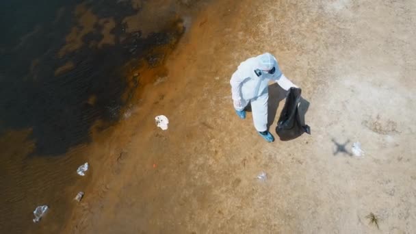 保護スーツを着た男と 乾燥した汚染された川のほとりにプラスチックゴミを集める人工呼吸器の空中写真 生態学的な大きなトロフィー人為的影響4Kドローン映像 — ストック動画
