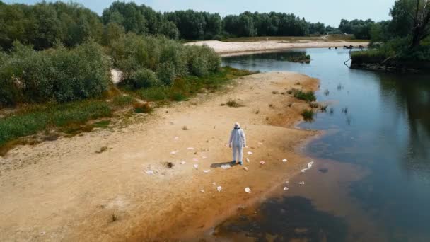 保護服を着た男と 乾燥した汚染された川の土手のプラスチック廃棄物の間に立っている人工呼吸器の空中写真 生態学的な大きなトロフィー人為的影響4Kドローン映像 — ストック動画