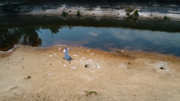 乾いた汚染された川のほとりにあるプラスチック製のゴミ箱の中でひざまずいている防護服と人工呼吸器を着た男の空中写真は ガスマスクを脱いで空気を吸う 4Kドローン映像 — ストック動画