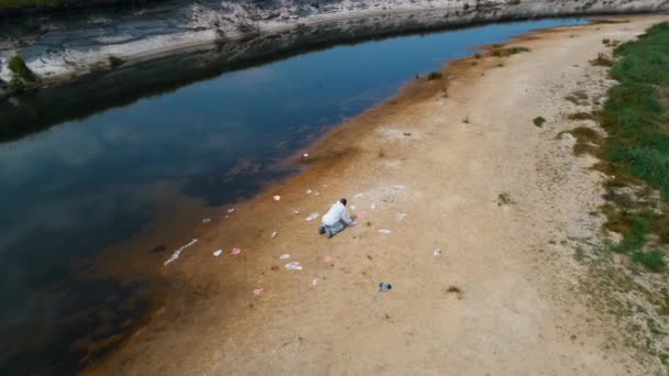 乾いた汚染された川のほとりにプラスチック製のゴミ箱の中にひざまずいている保護服を着た男の航空写真 生態学的な大きなトロフィー人為的影響4Kドローン映像 — ストック動画