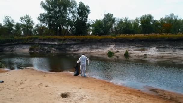 乾燥した汚染された川のほとりにプラスチックゴミを集める防護服を着た男の航空写真 生態学的な大きなトロフィー人為的影響4Kドローン映像 — ストック動画