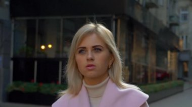 Pembe ceketli genç ve çekici sarışın bir kadın, modern mimarinin arka planına karşı duruyor. Güzel bir kız sokakta dururken düşünüyor. Yaşam tarzı, şehir, şehir. 4k görüntü.