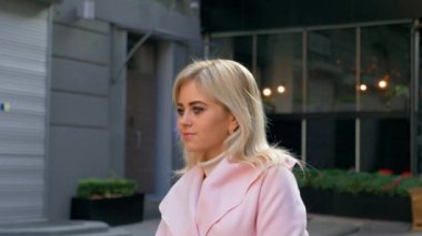 Pembe ceketli genç ve çekici sarışın bir kadın, modern mimarinin arka planına karşı duruyor. Güzel bir kız sokakta, gülümseyerek duruyor ve etrafa bakıyor. Yaşam tarzı, şehir, şehir. 4k