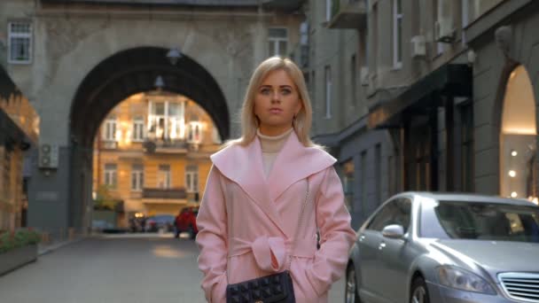 一位穿着粉红色外套的年轻迷人的金发女郎站在一条现代城市的街道上 美丽的女孩在站在汽车背景的户外思考 生活方式 — 图库视频影像