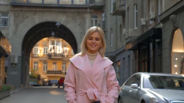 一位穿着粉红色外套的年轻迷人的金发女郎站在一条现代城市的街道上 美丽的女孩微笑着 而站在户外的汽车背景 生活方式 — 图库视频影像