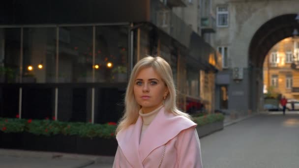 ピンクのコートを着た若い魅力的なブロンドの女性は 現代建築の背景に立っています 美しい少女が通りに立ってカメラを見る ライフスタイル 4Kフッテージ — ストック動画