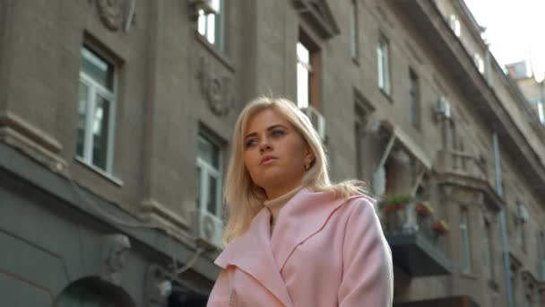 ピンクのコートを着た若い魅力的なブロンドの女性は 古い建築の背景に立っています 通りに立ちながら美しい少女が考えている ライフスタイル 4Kフッテージ — ストック動画