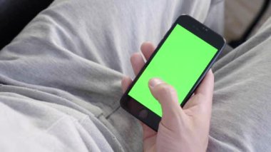 Adam evde kanepede oturan ve yeşil maket ekran ile akıllı telefon kullanarak. Adam kanepeye uzanmış, cep telefonuyla swiping yapıyor. Kaydırma hareketleri. Internet Sosyal Ağlar Tarama. Close. 4k