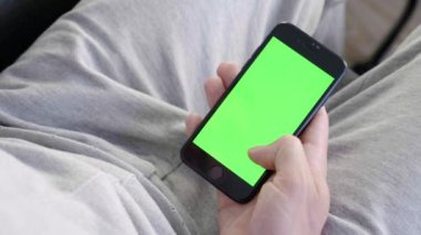 Adam evde kanepede oturan ve yeşil maket ekran ile akıllı telefon kullanarak. Adam kanepeye uzanmış, cep telefonuyla swiping yapıyor. Kaydırma hareketleri. Internet Sosyal Ağlar Tarama. Close. 4k