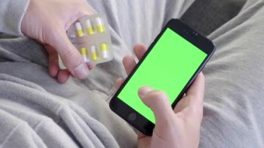 Adam yeşil maket ekran ile akıllı telefon kullanarak kanepede oturan ve hap alır. Adam cep telefonuyla swiping yapıyor. Kaydırma hareketleri. Internet Sosyal Ağlar Tarama. Hastalık soğuk algınlığı. Yakın çekim 4k