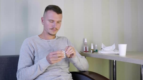 病人在家自食其人 一个心烦意乱的年轻人坐在沙发上 吃着药 喝着水 床头柜上有很多药片和药物 感冒病4K — 图库视频影像