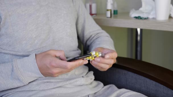 病気の男は家で自己投薬している スマートフォン上の検索エンジンで錠剤の情報を探している男 テーブルの上にたくさんの薬 急性呼吸器ウイルス感染風邪インフルエンザクローズ アップ — ストック動画