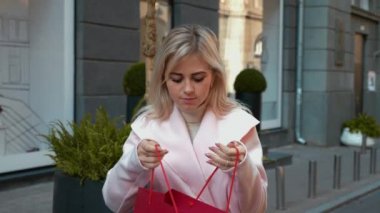 Pembe ceketli genç çekici sarışın kadın alışveriş. Bayan kırmızı bir kağıt torba içinde bir hediye ile mutlu, mutlulukla atlama. Noel tatilleri, satış ve perakende sektörü konsepti. 4k görüntü.