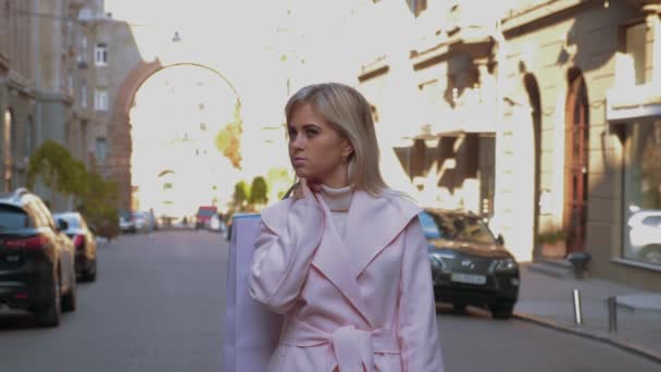 年轻迷人的金发女郎在粉红色的外套是购物 带着纸袋的女士站在汽车背景的大街上 面带微笑 圣诞假期 零售行业概念 — 图库视频影像