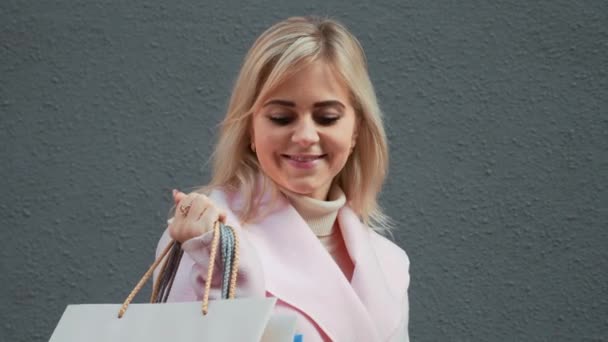 ピンクのコートを着た若い魅力的なブロンドの女性が買い物です 紙袋を持つ女性は 服を買った後 笑顔で楽しくジャンプしています クリスマス休暇 小売業界のコンセプト 4Kフッテージ — ストック動画