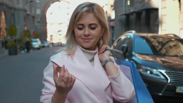 穿着粉红色外套的年轻迷人的女人正在购买加密货币 街上的女士拿着比特币的金币 向镜头展示 圣诞假期 零售行业概念 — 图库视频影像