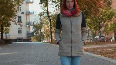 Ceketli, çekici, kızıl saçlı kadın cadde boyunca yürüyor. Güneşli bir günde dışarıda gezen seksi saçlı bir kız. Sonbahar mevsimi. Modern bir şehirde hayat. Hava güzel. 4k görüntü.