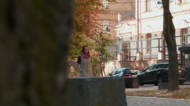 Ceketli, çekici, kızıl saçlı kadın cadde boyunca yürüyor. Güneşli bir günde dışarıda gezen seksi saçlı bir kız. Sonbahar mevsimi. Modern bir şehirde hayat. Hava güzel. 4k görüntü.