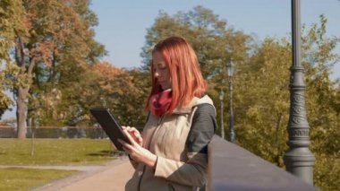 Parkta ceketli, çekici kızıl saçlı kadın internetten alışveriş yapıyor. Seksi saçlı bir kız açık havada güneşli havada durup tablet kullanıyor ve internetten sipariş veriyor. Satış, perakende sektörü konsepti. 4k