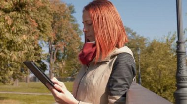 Parkta ceketli, çekici kızıl saçlı kadın internetten alışveriş yapıyor. Seksi saçlı bir kız açık havada güneşli havada durup tablet kullanıyor ve internetten sipariş veriyor. Satış, perakende sektörü konsepti. 4k