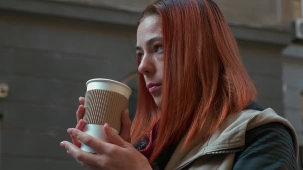 若い魅力的な赤い髪の女性ホットコーヒーを飲んでいます 路上の紙コップでセクシーな髪のお茶を飲む女の子 秋の寒い季節 現代都市での生活 都市生活 接近中だ4K映像 — ストック動画