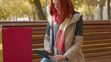 Parkta ceketli, çekici kızıl saçlı kadın internetten alışveriş yapıyor. Güneşli havada bankta oturan seksi saçlı kız tablet kullanıyor ve internetten sipariş veriyor. Satış, perakende sektörü konsepti. 4k
