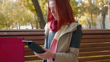 Parkta ceketli, çekici kızıl saçlı kadın internetten alışveriş yapıyor. Güneşli havada bankta oturan seksi saçlı kız tablet kullanıyor ve internetten sipariş veriyor. Satış, perakende sektörü konsepti. 4k