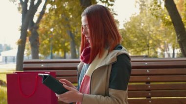 Parkta ceketli, çekici kızıl saçlı kadın internetten alışveriş yapıyor. Güneşli havada bankta oturan seksi saçlı kız tablet kullanıyor ve internetten sipariş veriyor. Satış, perakende sektörü konsepti. 4k