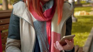 Parkta internetten alışveriş yapan çekici kızıl saçlı bir kadın. Seksi saçlı bir kız güneşli havada bankta oturur tablet ve telefon kullanır ve internetten sipariş verir. Satış, perakende sektörü konsepti. Yakın plan. 4k
