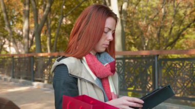 Parkta ceketli, çekici kızıl saçlı kadın internetten alışveriş yapıyor. Seksi saçlı bir kız açık havada güneşli havada durup tablet kullanıyor ve internetten sipariş veriyor. Satış, perakende sektörü konsepti. 4k