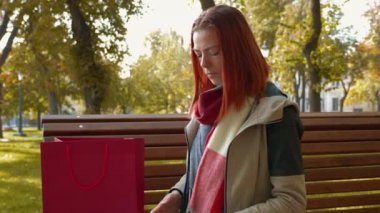 Parkta internetten alışveriş yapan çekici kızıl saçlı bir kadın. Seksi saçlı bir kız güneşli havada bankta oturur tablet ve telefon kullanır ve internetten sipariş verir. Satış, perakende sektörü konsepti. 4k görüntü.