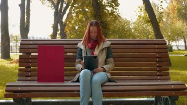 Parkta ceketli, çekici kızıl saçlı kadın internetten alışveriş yapıyor. Güneşli havada bankta oturan seksi saçlı kız tablet kullanıyor ve internetten sipariş veriyor. Satış, perakende sektörü konsepti. 4k
