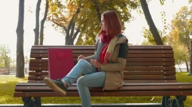 Parkta ceketli, çekici kızıl saçlı kadın internetten alışveriş yapıyor. Kürsüde oturan ve kırmızı bir kesekağıdından tablet alan seksi saçlı bir kız. Satış, perakende sektörü konsepti. 4k