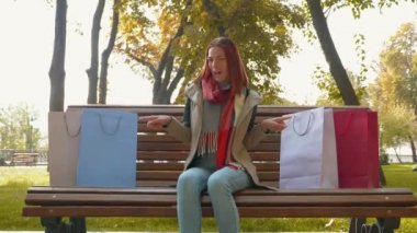 Genç ve çekici kızıl saçlı kadın alışveriş yapıyor. Parktaki bankta oturan seksi saçlı bir kız kağıt torbaları satın aldığına işaret ediyor. Satış, perakende sektörü konsepti. 4k görüntü.
