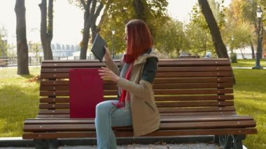 Parktaki çekici kızıl saçlı kadın internetten alışveriş yapıyor. Seksi saçlı bir kız bankta oturur ve kırmızı bir kesekağıdından tablet alıp kullanmaya başlar. Satış, perakende sektörü konsepti. 4k görüntü