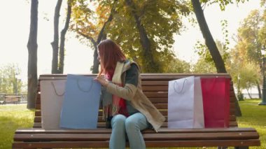 Genç ve çekici kızıl saçlı kadın alışveriş yapıyor. Parktaki bankta oturan ve alışverişini kağıt torbalarla yapan seksi saçlı bir kız. Satış, perakende sektörü konsepti. 4k