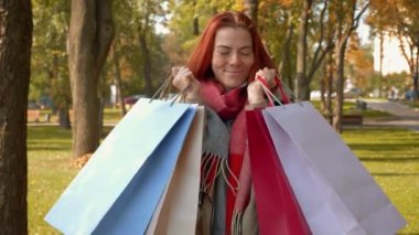 Ceketli, çekici, kızıl saçlı kadın alışveriş yapıyor. Parkta seksi saçlı mutlu bir kız çok renkli kese kağıtları içinde alışverişlerle ve sevinçle dolaşıyor. Satış, perakende sektörü konsepti. 4k görüntü.