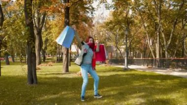 Ceketli, çekici, kızıl saçlı kadın alışveriş yapıyor. Parkta seksi saçlı mutlu bir kız çok renkli kese kağıtları içinde alışverişlerle ve sevinçle dolaşıyor. Satış, perakende sektörü konsepti. 4k görüntü.